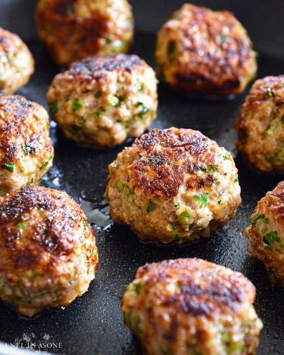 The image shows several round zucchini meatballs on a black baking tray, each about the size of a golf ball. The meatballs have a rough, uneven texture with visible bits of green zucchini and some browned spots, showing they are cooked. The color is mostly light brown with flecks of green and some darker areas where they browned more. The meatballs are spaced out, with one in the foreground clearly in focus and the others slightly blurred in the background. The scene is close-up and sharp on the front meatball, with soft lighting highlighting its texture. Photo taken with an iphone --ar 4:5 --v 7