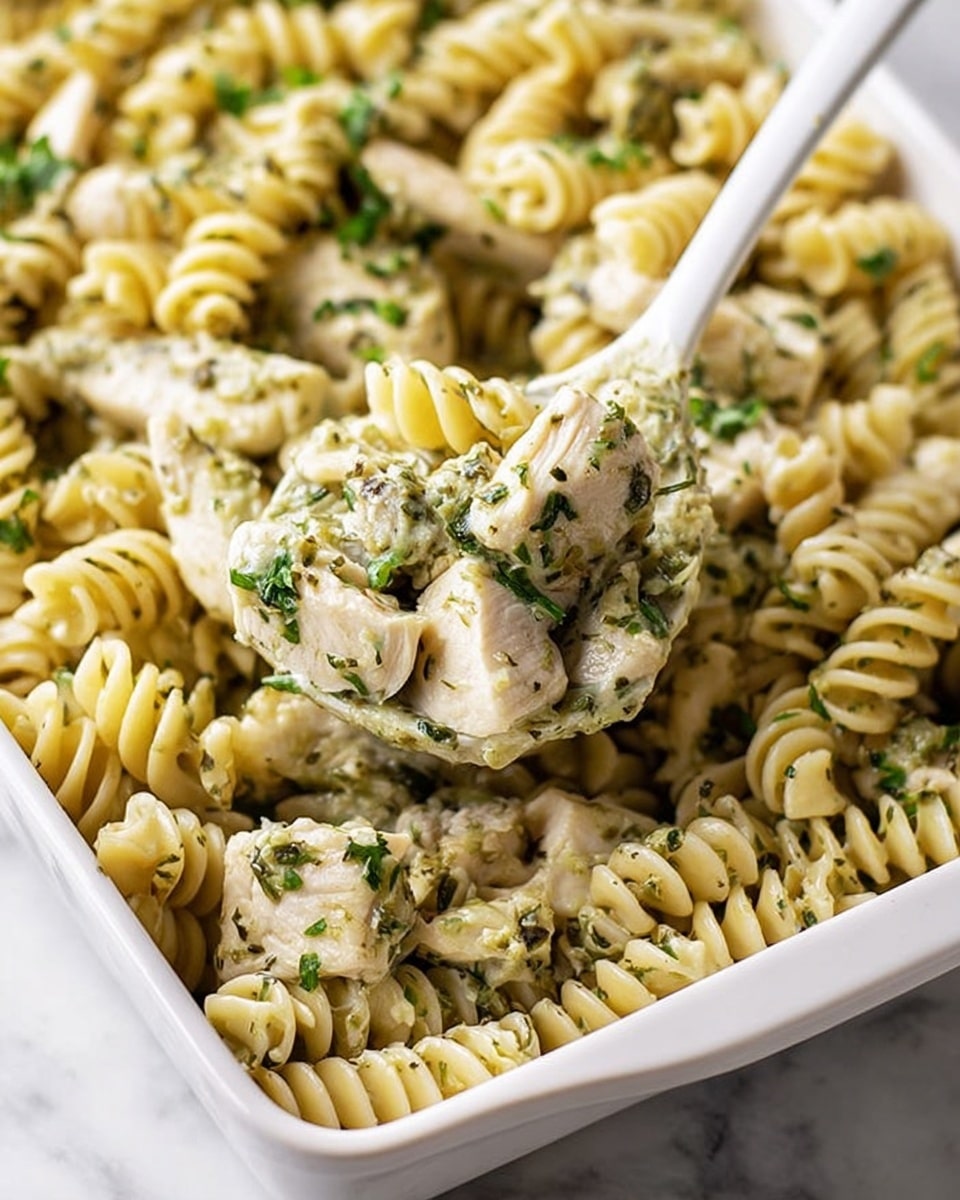 A white rectangular dish filled with creamy pasta made of light beige spiral rotini noodles mixed evenly with pieces of cooked white chicken breast, all coated in a green herb sauce with visible bits of parsley and other herbs, giving parts of the noodles a speckled green look; a white spoon is lifting a portion showing close-up details of the sauce and chicken pieces, against a white marbled background. photo taken with an iphone --ar 4:5 --v 7