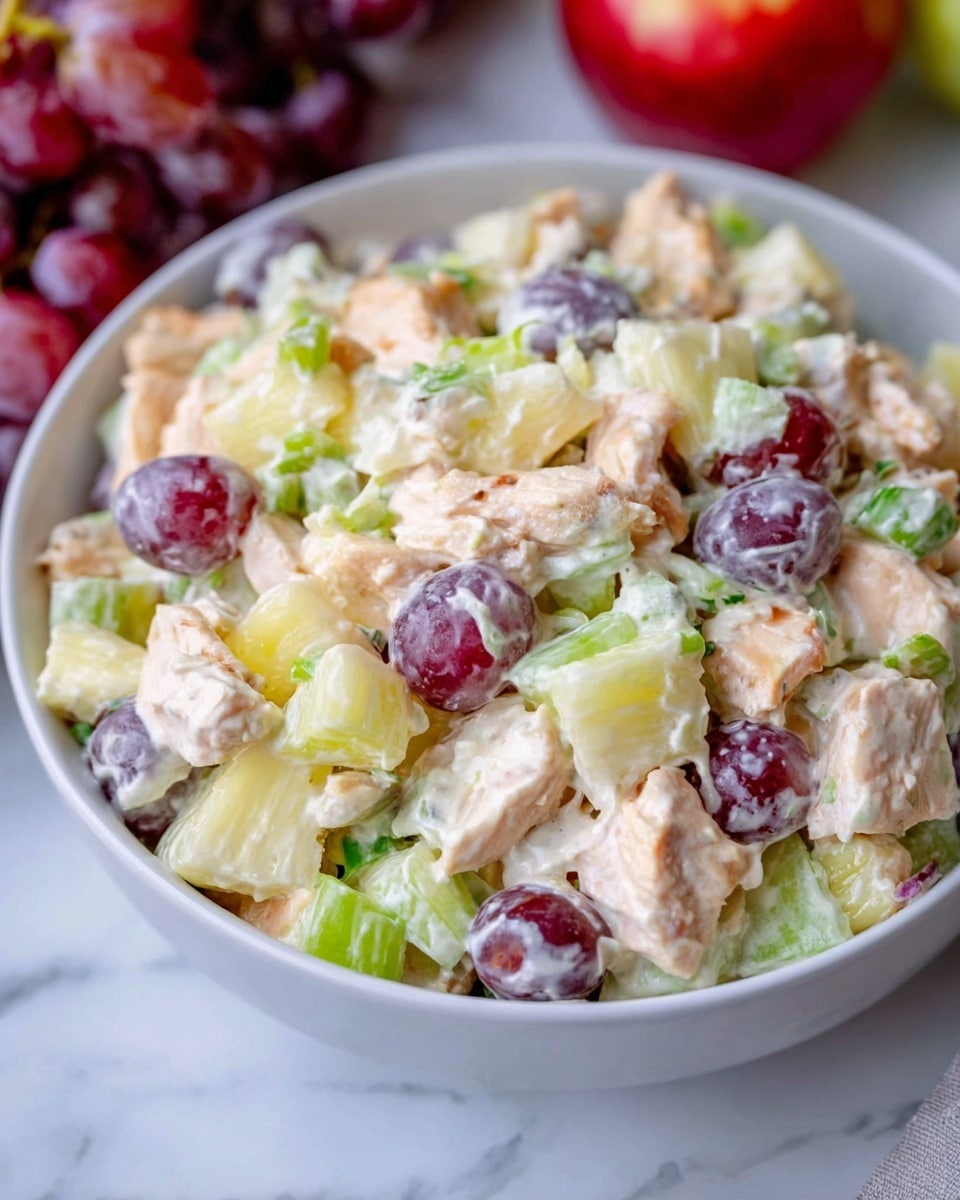 The image shows a white bowl filled with a creamy chicken salad. The salad has chunks of cooked chicken that are light tan in color, mixed with pale yellow pineapple pieces, bright green celery pieces, and dark purple halved grapes. The creamy dressing lightly coats each piece, giving a shiny, slightly wet look. The bowl is set on a white marbled surface with some red grapes and an apple blurred in the background. Photo taken with an iphone --ar 4:5 --v 7