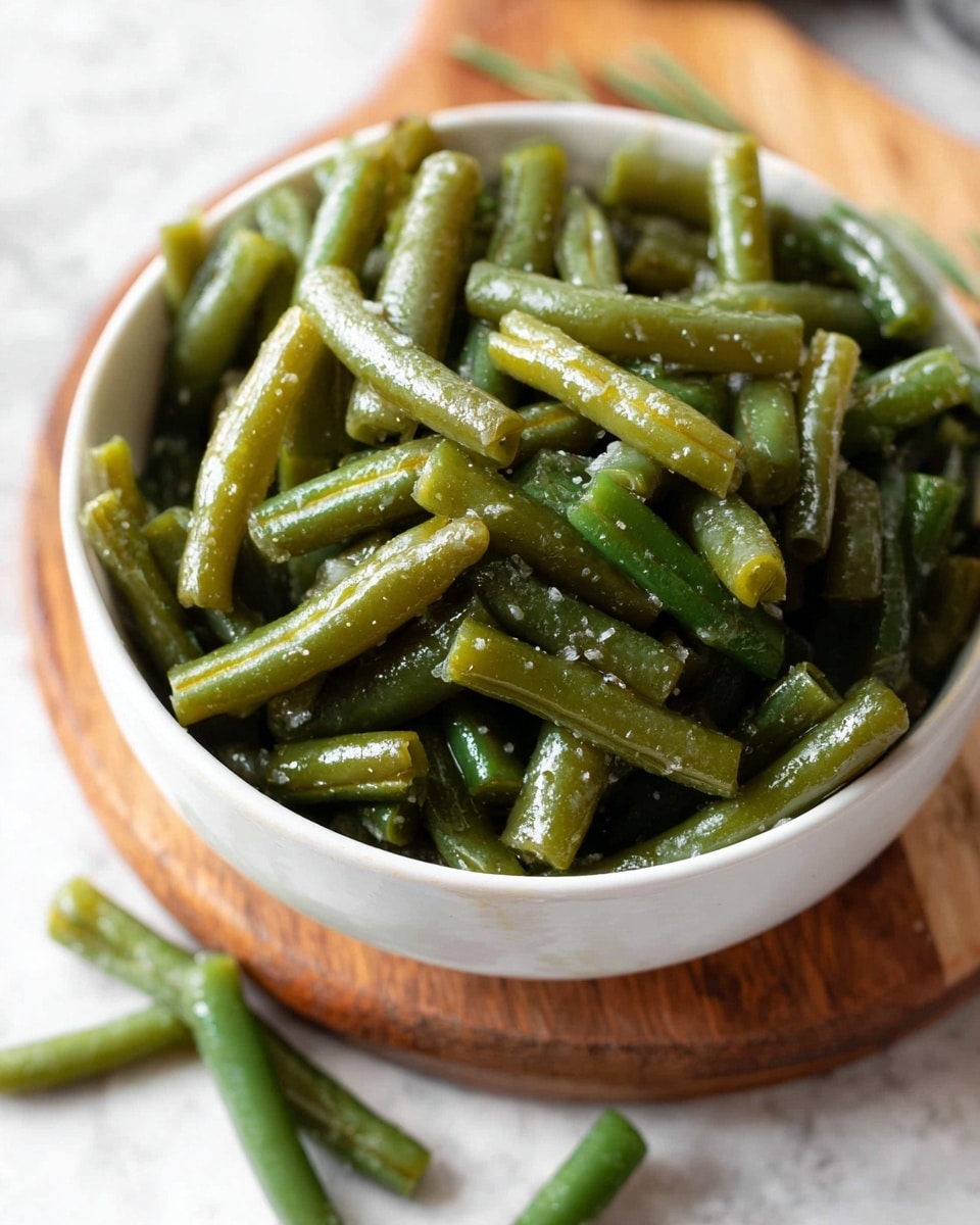 A close-up view of cooked green beans placed inside a white bowl, filled to the top with shiny, slightly oily green bean pieces that have a soft texture. The bowl is resting on a small wooden board, with a few green beans scattered around on the board and the white marbled surface beneath. The image shows the beans in varied angled positions, emphasizing their fresh, tender look and seasoning. photo taken with an iphone --ar 4:5 --v 7