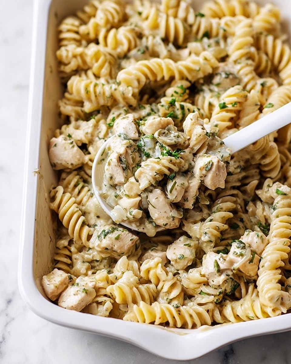 The image shows a white baking dish filled with creamy rotini pasta mixed with bite-sized pieces of cooked chicken. The pasta is light golden with a smooth texture, coated in a pale, herb-speckled sauce with visible green herbs and small bits of onion. The chicken pieces are tender and white, scattered evenly throughout the pasta. A white spoon is lifting a portion of the pasta and chicken, emphasizing the creamy texture and herbs. The whole dish sits on a white marbled surface that adds a clean and simple look. photo taken with an iphone --ar 4:5 --v 7