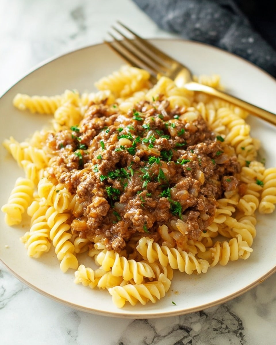 The image shows a white plate filled with three layers: at the bottom, there are yellow spiral rotini pasta pieces with a smooth texture, scattered unevenly around the edges; on top of the pasta, a thick layer of brown ground beef mixed with translucent cooked onion pieces creates a chunky and moist texture; sprinkled over the dish are small green chopped herbs evenly decorating the top, adding a fresh contrast. A gold fork is placed behind the food on the plate, and the whole scene is set against a white marbled surface. Photo taken with an iphone --ar 4:5 --v 7