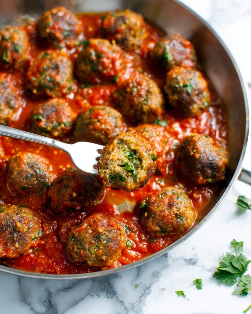 A close-up view of a metal pan filled with about fifteen small meatballs sitting in a thick, bright red tomato sauce with green herbs mixed in. The meatballs have a browned texture with bits of green visible inside them, showing they contain herbs or vegetables. In the pan, a metal fork is lifting one meatball, highlighting the juicy and moist texture. The background is a white marbled surface with a few scattered herb leaves around the pan photo taken with an iphone --ar 4:5 --v 7