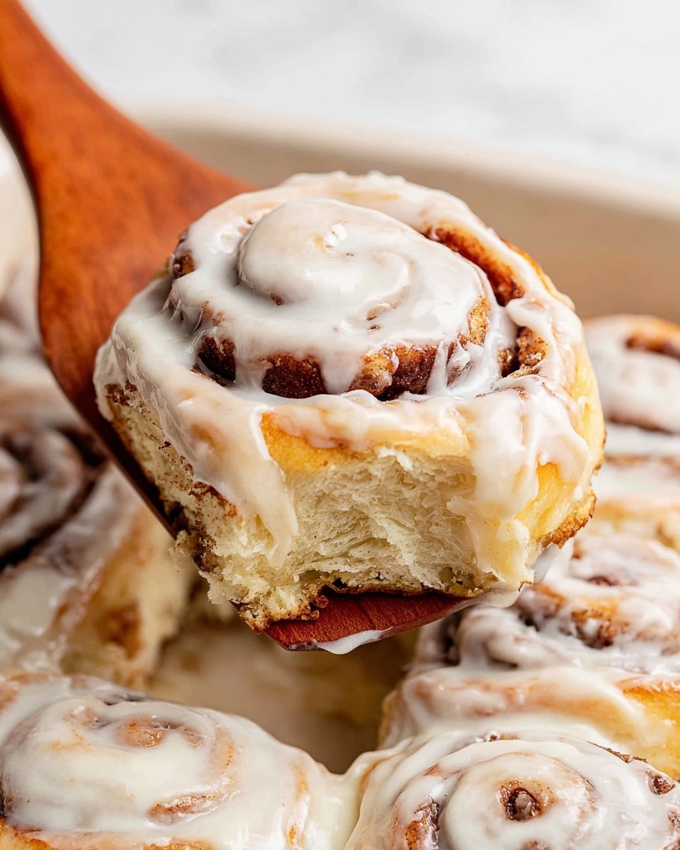 The image shows a close-up of four cinnamon rolls in a white baking dish, all covered with thick white cream cheese icing. Each roll has a golden brown color with visible swirls of cinnamon inside. The icing looks smooth, creamy, and slightly melted, spreading over the top and down the sides of the rolls. The white baking dish holds the rolls tightly together, showing soft, fluffy texture on the edges. The background is a white marbled texture. photo taken with an iphone --ar 4:5 --v 7