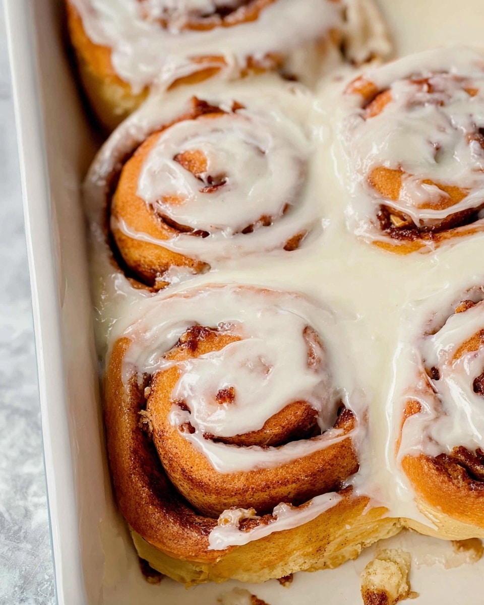 A close-up view of a cinnamon roll with three visible layers: the bottom layer is a soft, light golden dough, the middle layer shows dark brown cinnamon filling rolled in a spiral shape, and the top layer is thick white icing that slightly drips down the sides. The cinnamon roll is held by a wooden spatula above a white baking dish filled with more cinnamon rolls covered with the same creamy white icing. The background is a white marbled texture. photo taken with an iphone --ar 4:5 --v 7