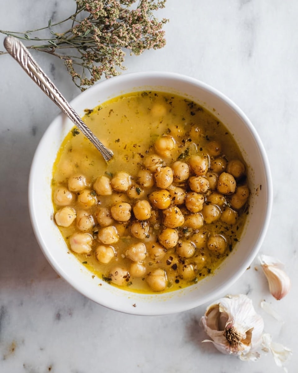 A white bowl filled with a warm chickpea stew, showing two layers: the top layer has light brown chickpeas mixed in a yellowish broth with visible small dark pepper specks and herbs, while the bottom layer is a clearer golden broth. Inside the bowl, a silver spoon with a twisted handle rests on the left side, slightly dipped into the stew. The bowl is placed on a white marbled surface, with a sprig of dried small flowers at the top and a garlic bulb broken into cloves at the bottom right corner, adding a rustic touch. Photo taken with an iphone --ar 4:5 --v 7