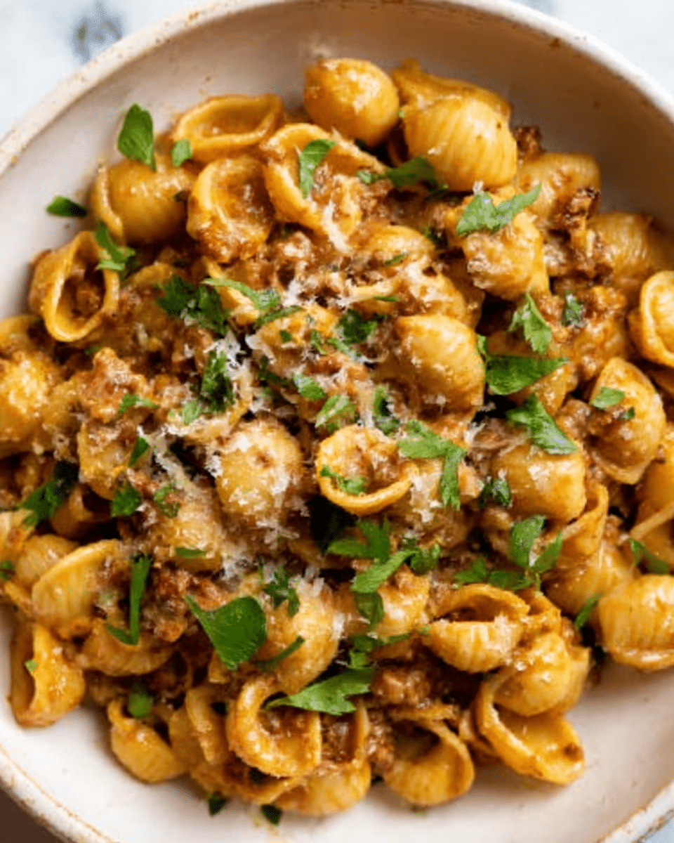 The image shows a white bowl filled with small pasta shells coated in a creamy brown sauce with visible bits of ground meat mixed throughout. Fresh green parsley leaves are scattered on top for color, and a light sprinkle of grated cheese dusts the surface. The pasta looks tender and saucy, with textures ranging from smooth sauce to slightly grainy meat and leafy herbs. The bowl sits on a white marbled surface. photo taken with an iphone --ar 4:5 --v 7