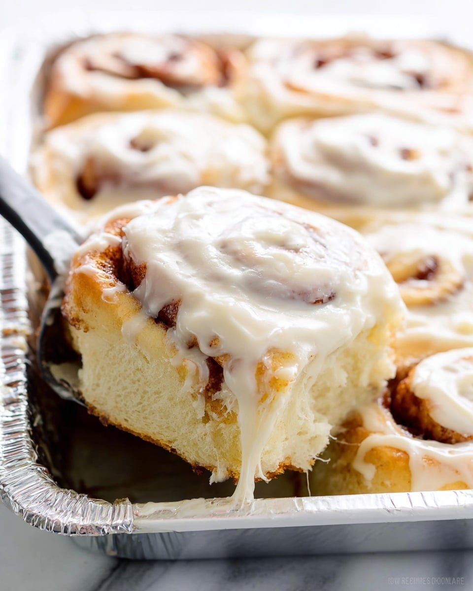 This image shows a close-up of a soft cinnamon roll being lifted from a white rectangular pan covered with foil. The cinnamon roll has three main layers: the bottom is a light golden-brown fluffy dough, the middle has swirled darker cinnamon filling, and the top layer is thick, smooth, and creamy white frosting spread generously and melting slightly over the roll’s edges. The texture looks light and moist with some strands of dough stretching as the roll is lifted, all sitting against a white marbled surface. photo taken with an iphone --ar 4:5 --v 7