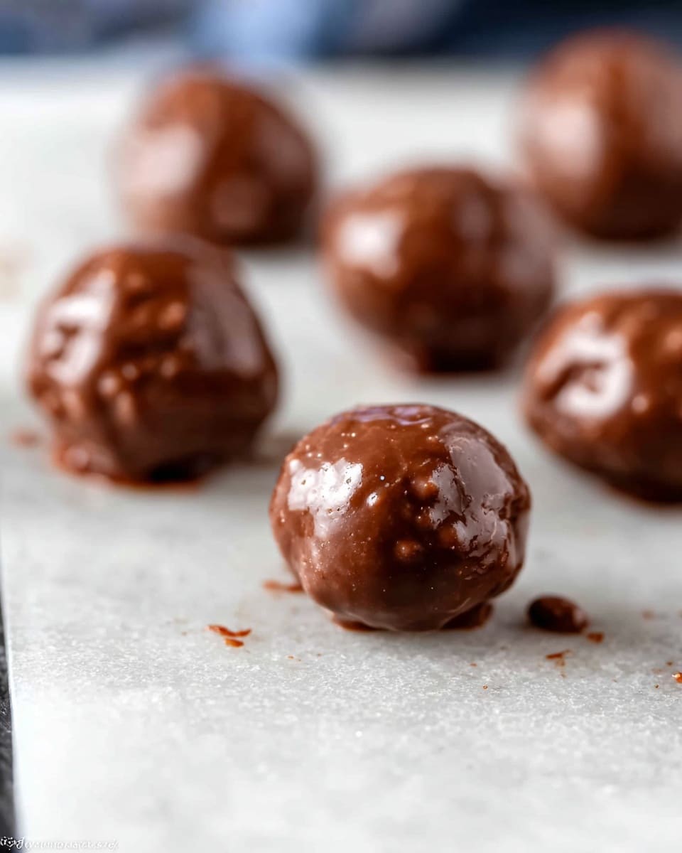 A group of round chocolate-coated treats arranged on a light-colored baking mat with a grid pattern, one treat in the foreground shows a bite taken out, revealing a crumbly, light brown inside. The chocolate coating is smooth and shiny, brown in color and covers the entire outside of each ball. There are small chocolate crumbs scattered around the treats on the mat, adding texture to the scene. The background is softly blurred, focusing attention on the front bitten chocolate ball. photo taken with an iphone --ar 4:5 --v 7
