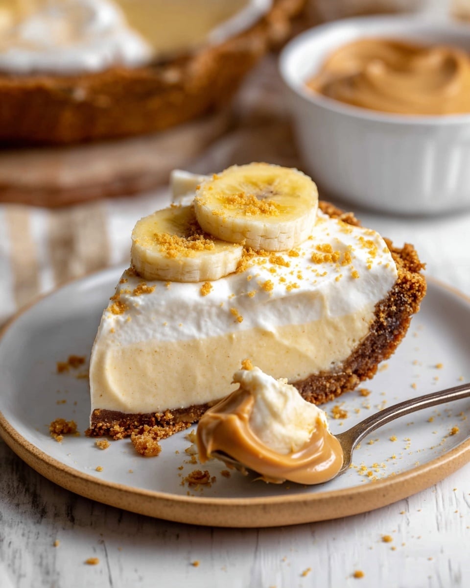 A slice of banana cream pie sits on a white plate with a light brown rim, placed on a white marbled surface. The pie has three clear layers: a thick, crumbly dark brown crust, a pale yellow creamy banana filling, and a fluffy white whipped topping sprinkled with small golden-brown cookie crumbs. A slice of banana with a light golden-brown caramelized edge rests on the whipped cream. A spoon on the plate holds a scoop of the pie showing the creamy texture of the filling and whipped topping. In the background, there is a blurred white bowl filled with smooth peanut butter. Photo taken with an iphone --ar 4:5 --v 7