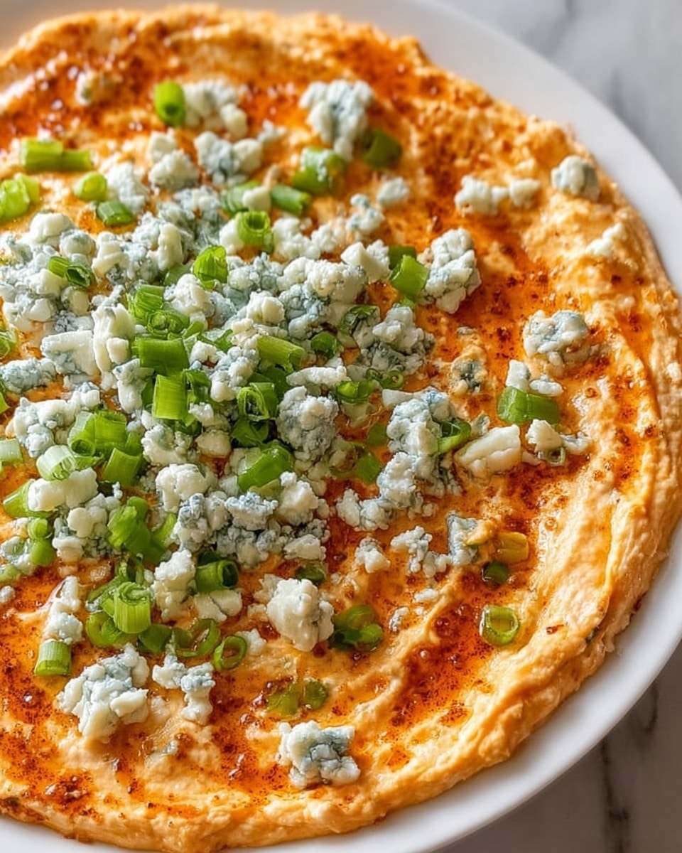 The image shows a close-up view of a creamy dip spread evenly on a white plate, creating a single smooth layer with an orange hue mixed with light cream tones. On top, there are crumbles of blue cheese scattered around the center, showing white and bluish-green textures. Diced green onions are sprinkled over the dip, adding spots of bright green color and a fresh texture contrast. The edge of the plate is cleanly visible, and the background is a white marbled surface. photo taken with an iphone --ar 4:5 --v 7