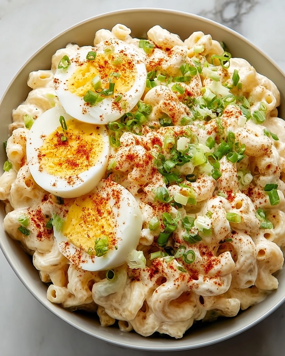 A white bowl filled with creamy, white macaroni salad made of small pasta shells, topped with three halves of bright yellow hard-boiled eggs sprinkled with red paprika powder. Fresh green chopped scallions are scattered on top, adding a touch of color and freshness. The dish sits on a white marbled surface, showing a rich, smooth texture of the sauce coating the pasta and garnishes. photo taken with an iphone --ar 4:5 --v 7