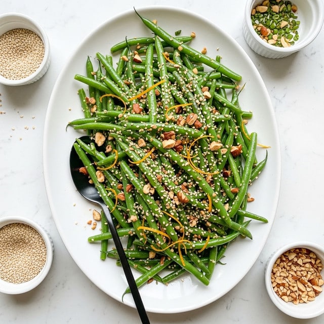 The dish shows a large white oval plate filled with bright green cooked green beans, tossed with sesame seeds and chopped nuts scattered on top, adding texture and contrast. Thin strips of orange zest are sprinkled across the green beans, giving a pop of color. A black spoon rests on the plate, partially covered by the green beans and nuts. Around the plate, there are small white bowls, one filled with sesame seeds, and another with chopped nuts, placed on a white marbled surface. The overall look is fresh and vibrant, with a mix of green, white, light brown, and orange colors. photo taken with an iphone --ar 4:5 --v 7