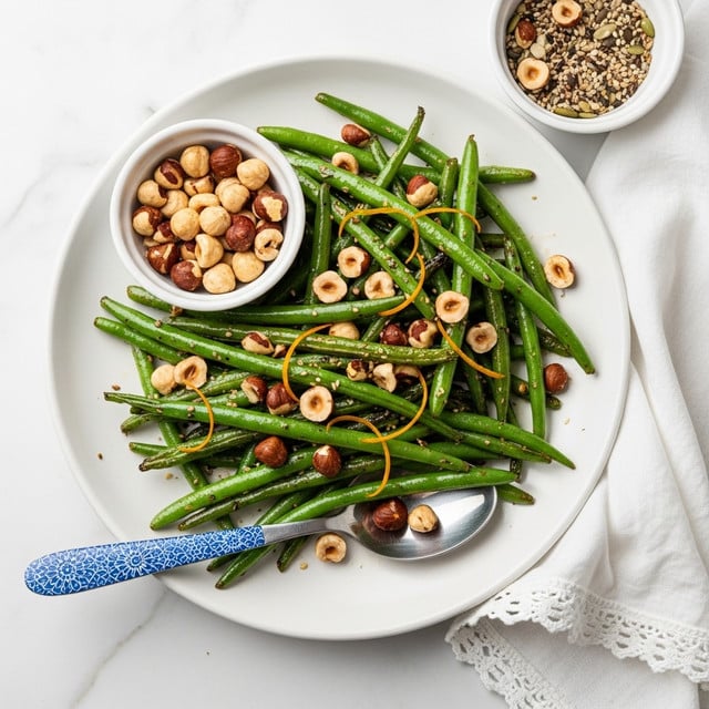 The image shows a white plate filled with a pile of cooked green beans that are glossy and slightly charred, topped with small whole and chopped hazelnuts and thin strips of orange zest. A small white bowl placed on the plate contains more whole and chopped hazelnuts. Next to the plate is another small bowl with a mix of seeds and nuts. A blue patterned spoon rests on the edge of the plate beneath some green beans. The plate and bowls sit on a white marbled surface with a white cloth featuring lace trim beside them. photo taken with an iphone --ar 4:5 --v 7