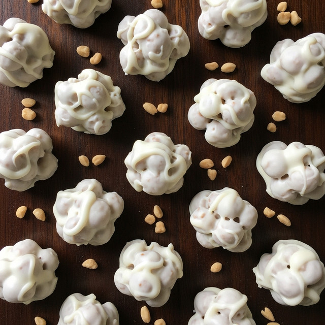 The image shows many small clusters of snack-sized treats covered in smooth, white coating with a slightly glossy finish, each with uneven, round shapes and visible lumps underneath the coating indicating nuts or other crunchy bits. These white clusters rest directly on a dark brown wooden surface, contrasting with the white treats. Scattered around the clusters are small pieces of light brown nuts adding texture and color contrast. The arrangement is evenly spaced, filling the entire frame with a top-down view. photo taken with an iphone --ar 4:5 --v 7