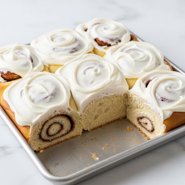 A close-up of a square cinnamon roll base with a thick layer of white creamy frosting spread unevenly on top, showing swirls and small peaks. The cinnamon roll has a golden brown outer crust with visible soft inner layers and cinnamon specks, and a corner piece is missing from the front right side, revealing the light fluffy texture inside. The roll is placed on a metal baking tray with some crumbs and baked spots visible around it, all set on a white marbled texture surface. Photo taken with an iphone --ar 4:5 --v 7