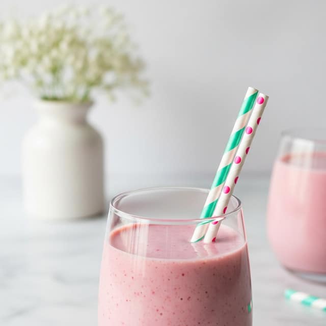 The image shows a close-up of a clear glass filled with a pink smoothie. Inside the glass, there are two paper straws, one striped in teal and white, and the other with pink dots on white. The glass sits on a white marbled surface. In the softly blurred background, there is a small white vase holding a bunch of white flowers, and another similar glass with a pink smoothie and a straw is partly visible on the right side. The colors are soft and pastel, creating a calm and fresh feel. Photo taken with an iphone --ar 4:5 --v 7