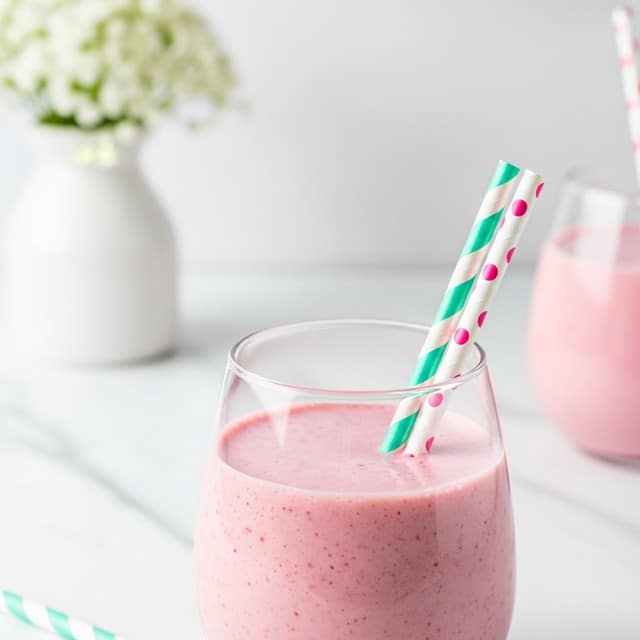 The image shows a clear glass filled with a light pink smoothie, about three-quarters full, with two paper straws sticking out from the top—one straw is white with pink dots, and the other is white with teal stripes. Behind the glass, there is a blurred white flower bouquet in a transparent vase set on a white marbled surface, and another glass with the same pink smoothie and a light pink striped straw is placed to the right, slightly out of focus. The background is a soft, pale pink color, adding to the gentle and fresh look of the scene. Photo taken with an iphone --ar 4:5 --v 7