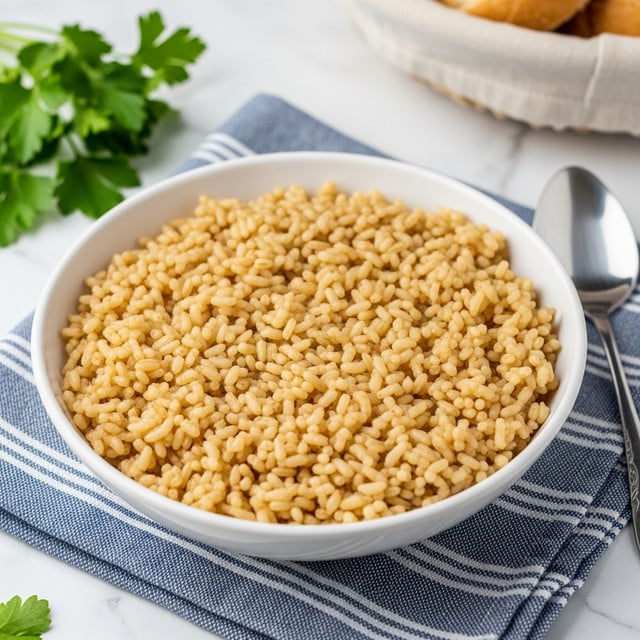 A close-up of a white bowl filled with cooked rice that is brown and slightly glossy, showing individual grains mixed with small browned pieces of a cooked ingredient, likely toasted vermicelli or spices. The rice looks soft and fluffy, with a textured surface. The bowl rests on a white marbled surface with a blue and white striped cloth partially visible under the bowl, fresh green parsley leaves on the left side, and a metal spoon with a long handle beside the bowl. A basket with light brown bread is blurred in the background. Photo taken with an iphone --ar 4:5 --v 7