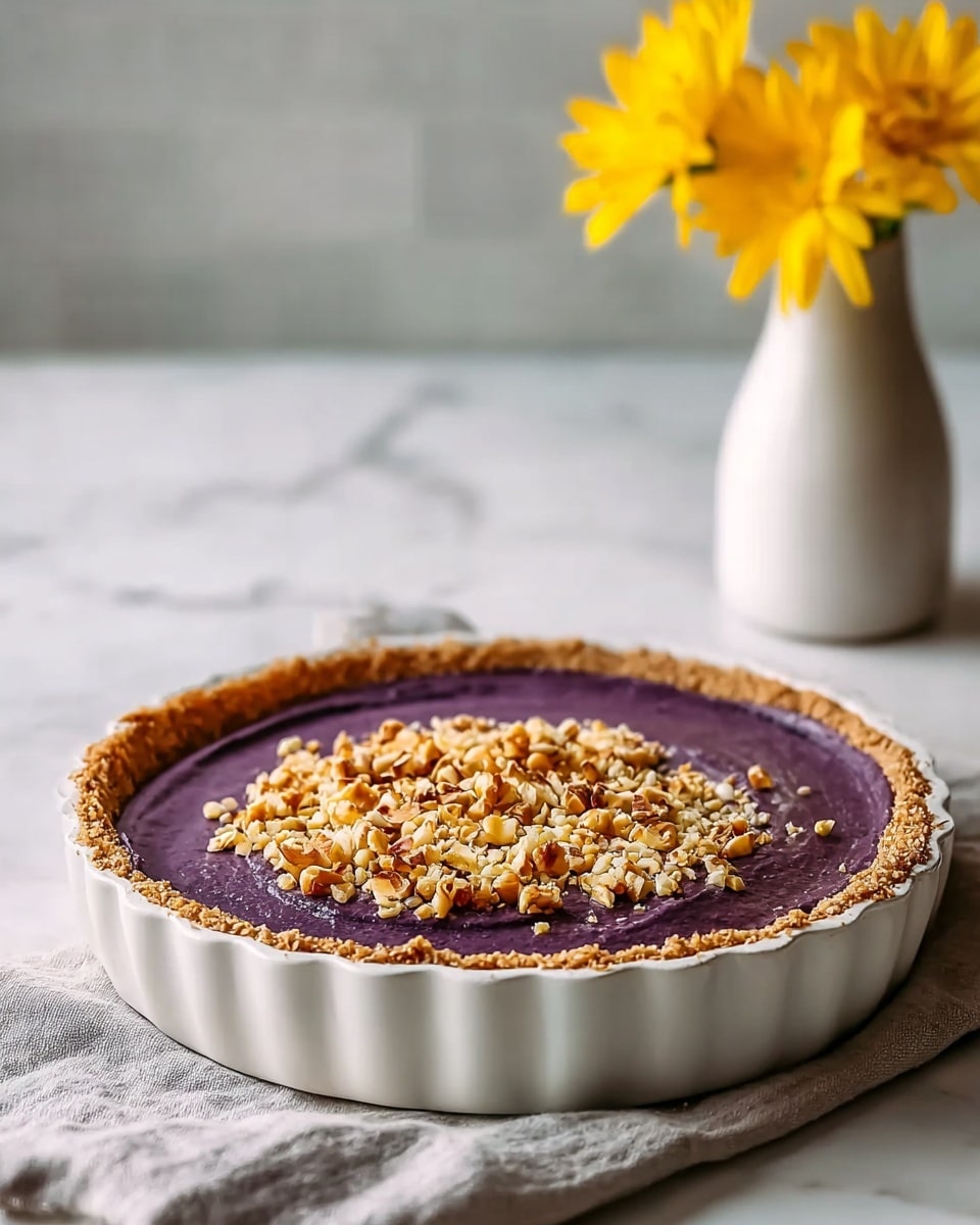 This image shows a pie with three visual layers in a white fluted pie dish. The bottom layer is a golden-brown crumbly crust forming the base and edges. The middle layer is thick and smooth, with a rich purple color filling most of the pie. The top layer is a textured sprinkle of light brown chopped nuts and crumbs, covering the center of the purple filling but leaving the edges clear to show the crust. The pie rests on a soft gray cloth, with some crumbs scattered around, and in the background is a simple white vase holding bright yellow flowers, all placed on a white marbled surface. Photo taken with an iphone --ar 4:5 --v 7