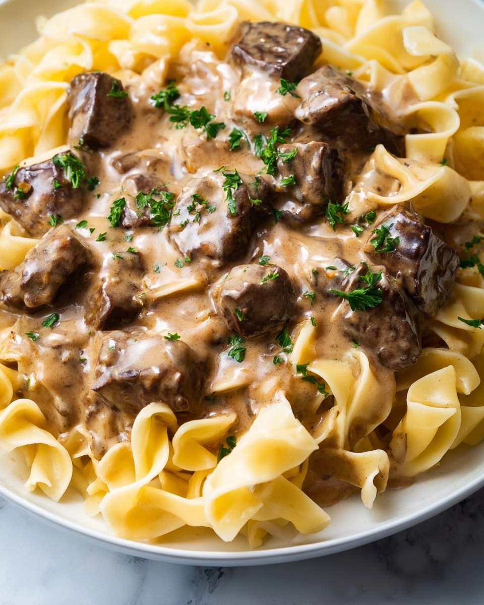 A close-up view of a dish featuring thick egg noodles at the base, soft and twisted with a pale yellow color and smooth texture. On top of the noodles are large, square and irregular chunks of beef covered in a rich, creamy brown sauce that coats the meat and noodles evenly. Small pieces of green parsley are scattered lightly over the sauce, adding a touch of color. The image is focused tightly on the food, showing the glossy and creamy texture of the sauce mingling with the tender beef and soft noodles. The background is a white marbled texture. photo taken with an iphone --ar 4:5 --v 7