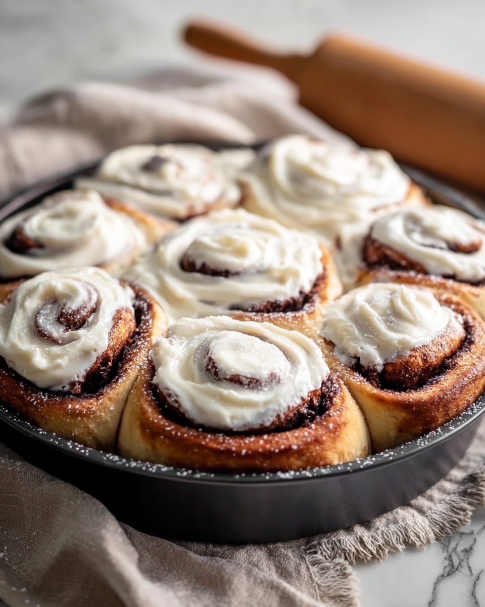A close-up of a round black pan filled with seven thick cinnamon rolls topped with creamy white icing. Each roll has three to four visible swirled layers, showing golden brown dough with darker cinnamon filling spiraled inside. The icing is generously spread on top, covering most of each swirl with a smooth and slightly glossy texture. There’s a light dusting of powdered sugar over the icing. The pan sits on a beige cloth on a white marbled surface with a wooden rolling pin in the background. Photo taken with an iphone --ar 4:5 --v 7