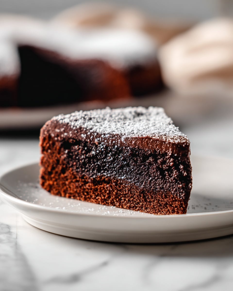 A single slice of rich chocolate cake sits on a white plate, showing two dense, moist layers of dark chocolate cake with a thick, creamy, chocolate filling in the middle. The top of the slice is dusted lightly with powdered sugar, adding a soft, snowy texture contrasting the dark cake. The plate rests on a white marbled surface with a blurred background containing another cake and some green foliage. photo taken with an iphone --ar 4:5 --v 7