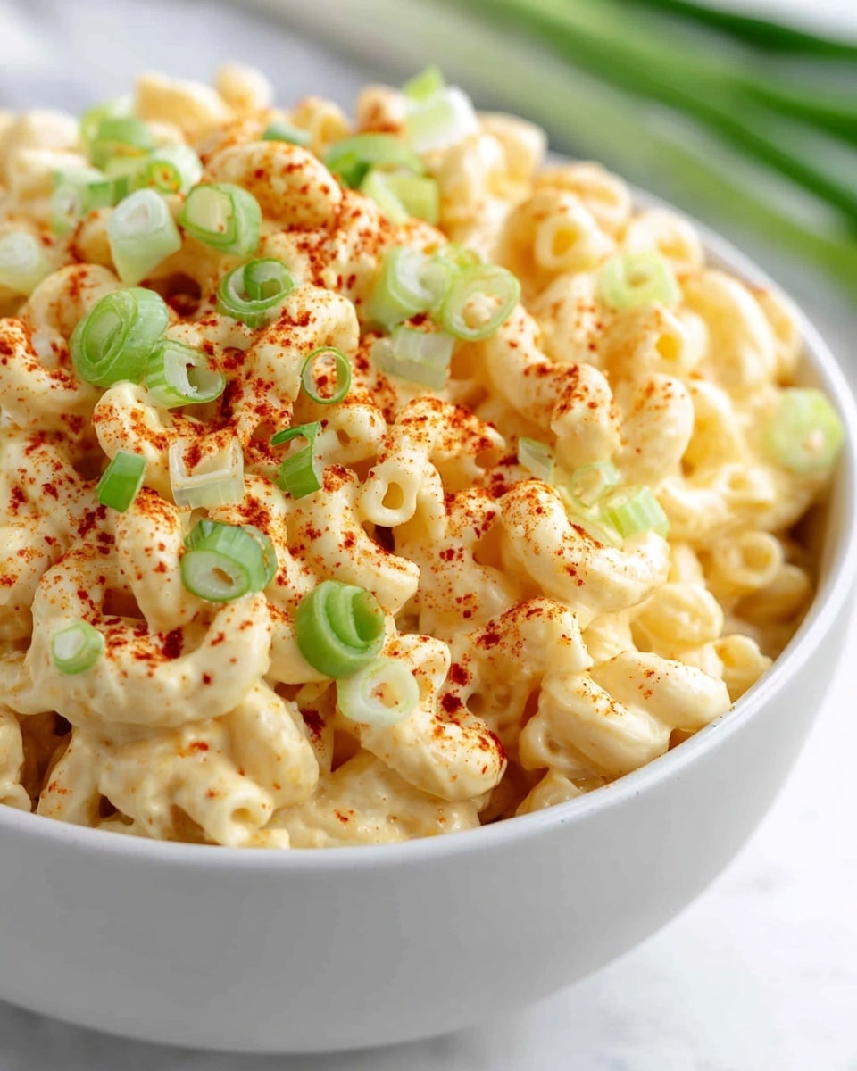A close-up view of a white bowl filled with curly macaroni pasta coated in creamy, light yellow cheese sauce. The top layer is sprinkled with bright red paprika powder and chopped fresh green onions, adding color contrast and texture. The bowl is resting on a white marbled surface, with blurred green onions in the background. The macaroni looks soft and well mixed with the cheese sauce, making it look rich and appetizing. Photo taken with an iphone --ar 4:5 --v 7