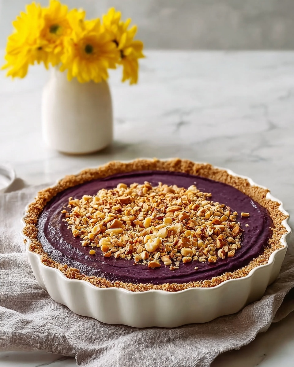 A pie with three layers is shown: the bottom layer is a crumbly, light brown crust that lines a white fluted pie dish, followed by a smooth, thick layer of deep purple filling that almost reaches the crust’s edge, and the top layer is a generous sprinkle of chopped nuts in various golden brown tones covering the center of the purple filling. The pie sits on a soft, light gray cloth on a white marbled surface. In the blurred background, there is a simple white vase holding bright yellow flowers. Photo taken with an iphone --ar 4:5 --v 7