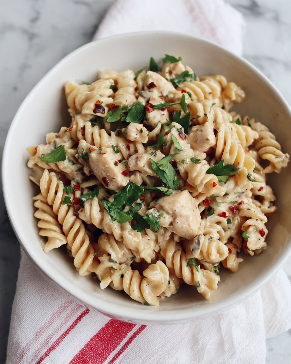 A white bowl filled with creamy rotini pasta mixed with pieces of cooked chicken. The pasta and chicken are coated in a light beige sauce with visible herbs and small red pepper flakes. Bright green parsley leaves are scattered on top, adding a fresh touch of color. The bowl is placed on a white cloth with a red stripe, set on a white marbled textured surface. photo taken with an iphone --ar 4:5 --v 7