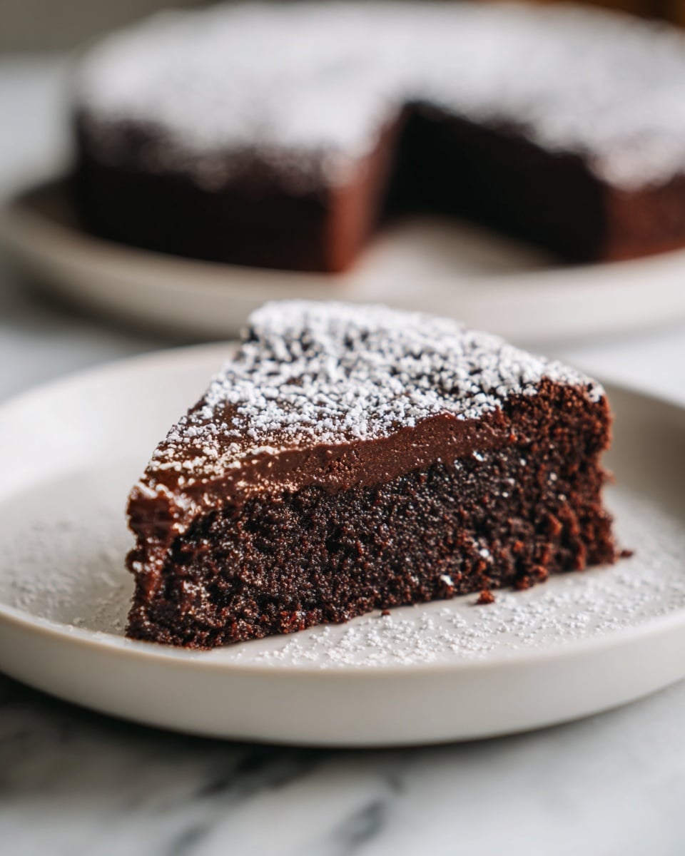 A close-up of a single slice of chocolate cake on a white plate, placed on a white marbled surface. The cake slice has two visible layers: a dense, moist dark chocolate base and a rich, slightly glossy chocolate topping. The surface of the cake is topped with a light dusting of white powdered sugar that contrasts with the deep brown color. In the background, there is a blurry whole chocolate cake. The photo is bright with natural light and soft shadows, showing the cake’s texture clearly. Photo taken with an iphone --ar 4:5 --v 7