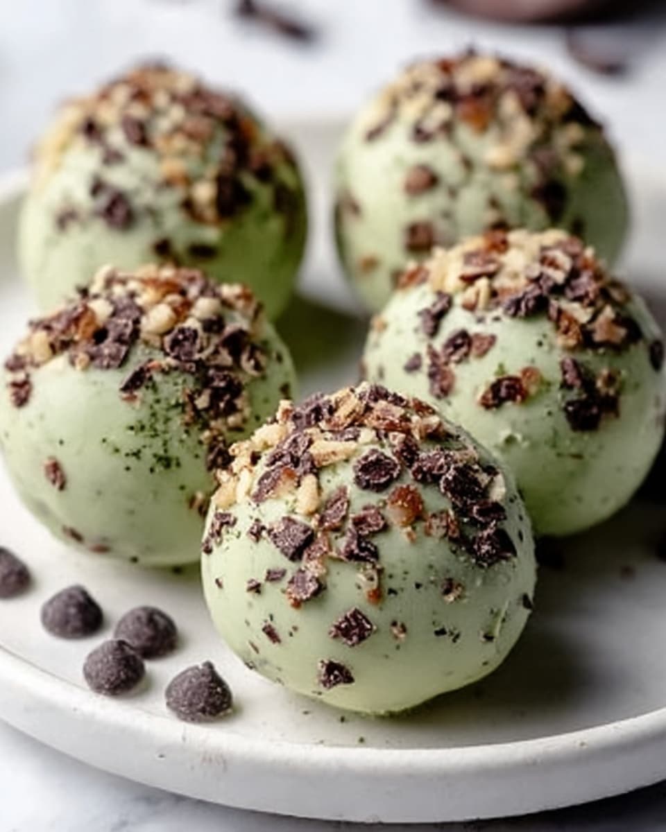 A close-up image of six round green truffles covered in crushed nuts and chocolate chips, resting on a white plate with extra chocolate chips scattered around the base. Each truffle appears smooth and glossy with a light green color, sprinkled evenly with small brown nut pieces and dark chocolate chunks, creating a crunchy texture on the surface. The scene is set on a white marbled surface, enhancing the contrast between the truffles and their surroundings. photo taken with an iphone --ar 4:5 --v 7