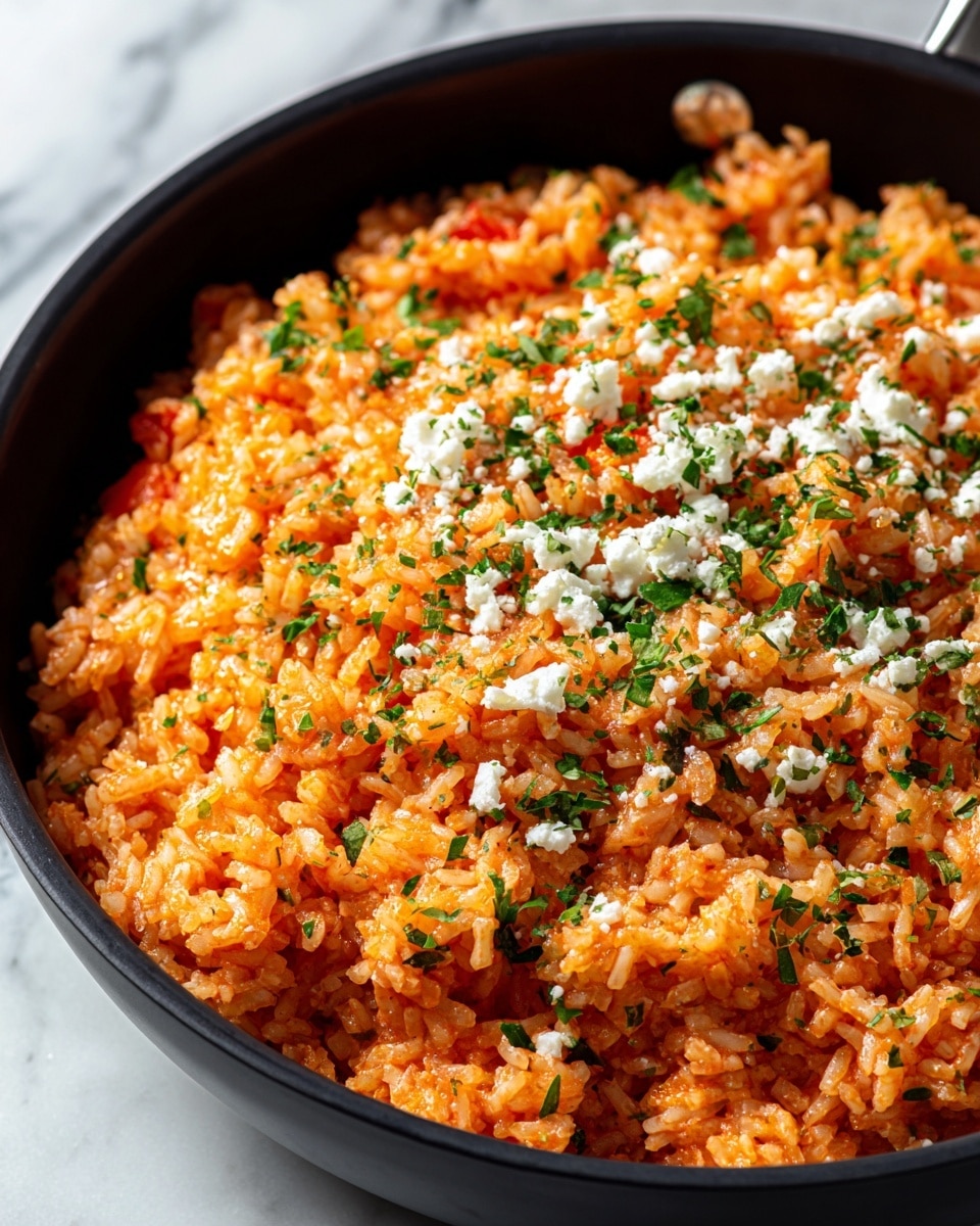 A close-up of a black pan filled with one layer of cooked rice mixed with finely chopped green herbs and small red bits, giving it an orange tint. The rice is sprinkled evenly with small white crumbly cheese on top, adding texture. The grains are separate but slightly sticky, showing a moist, fluffy texture under warm lighting. The pan is set on a white marbled surface. photo taken with an iphone --ar 4:5 --v 7