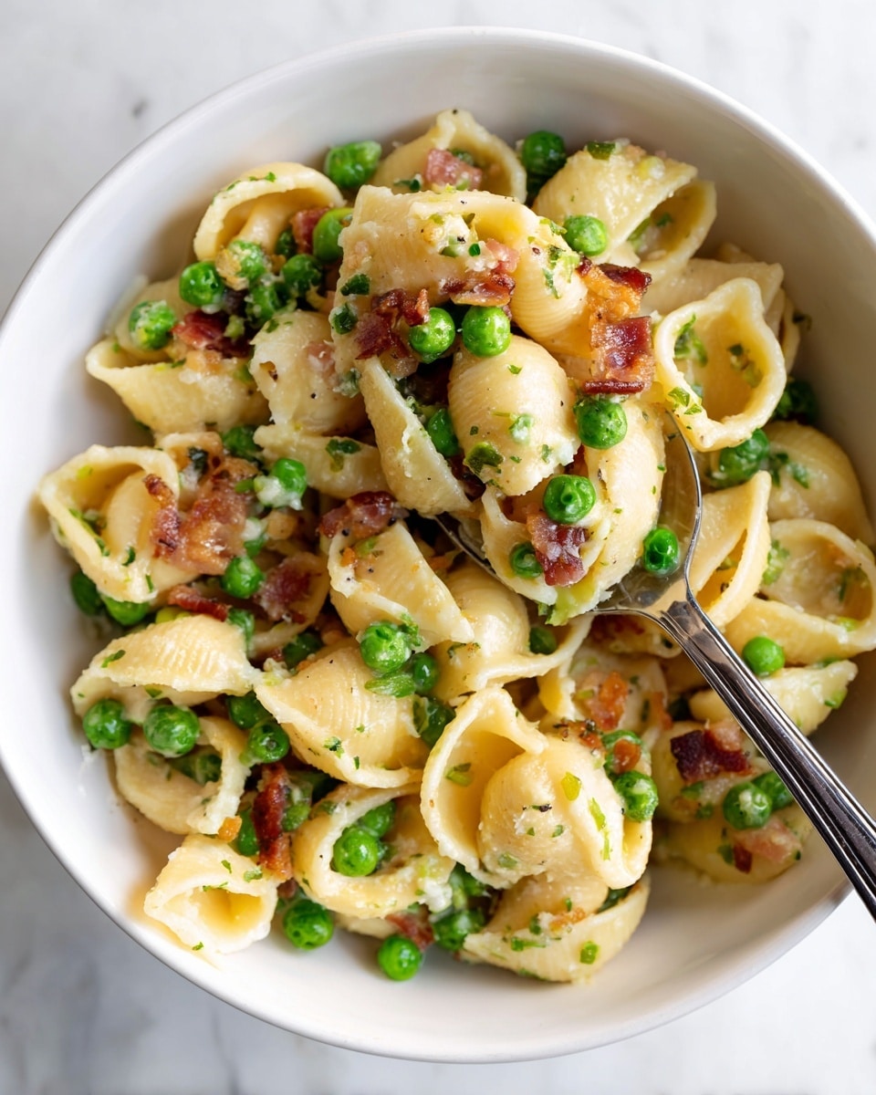 A close-up view of shell pasta mixed with green peas and small brown bacon bits, coated in a creamy light-colored sauce. The pasta shells are large and curved with ridges, and the green peas are bright and round, scattered evenly throughout. The bacon pieces add a crunchy texture, with a slightly crispy, reddish-brown appearance. A silver spoon holds a portion of the mix above a white bowl filled with the pasta dish. The background shows a white marbled surface. Photo taken with an iphone --ar 4:5 --v 7