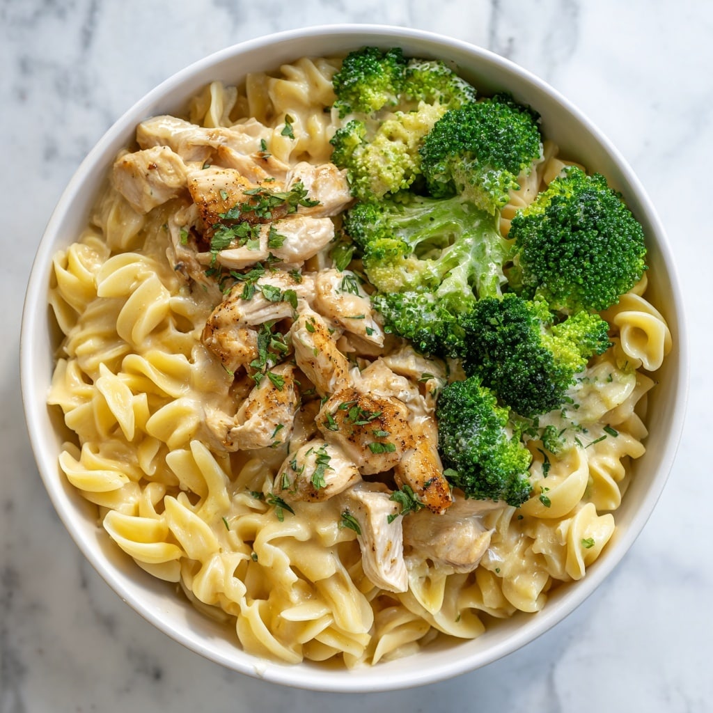 The image shows a bowl filled with a creamy pasta dish. It has three main layers: the bottom layer is small twisted pasta with a smooth, light yellow sauce, the middle layer contains bright green broccoli florets scattered around, and the top layer features pieces of cooked chicken that are light brown with a slight golden color. Tiny bits of fresh green herbs are sprinkled over the dish, adding texture and color. The bowl is white and placed on a white marbled surface. Photo taken with an iphone --ar 4:5 --v 7
