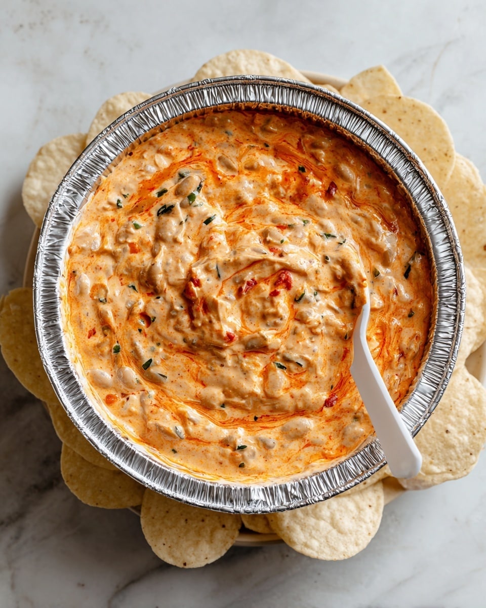 The image shows a single-layer creamy dip in a round aluminum foil container, with a thick, swirled texture mixing orange-red and white colors. The dip has some visible chunks of beans and small green herb pieces scattered throughout. The container sits on a white marbled surface surrounded by light beige round tortilla chips. A white plastic spoon is placed inside the dip, partially submerged. The photo taken with an iphone --ar 4:5 --v 7