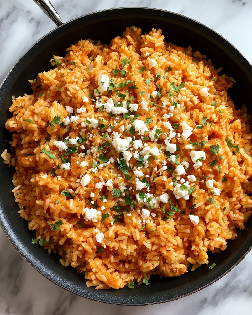 A close-up view of a pan filled with cooked rice mixed with tomato sauce giving it an orange color and moist texture, with tiny green herb pieces scattered throughout, lightly topped with crumbled white cheese that contrasts against the orange rice. The pan is black and the background is a white marbled texture. photo taken with an iphone --ar 4:5 --v 7