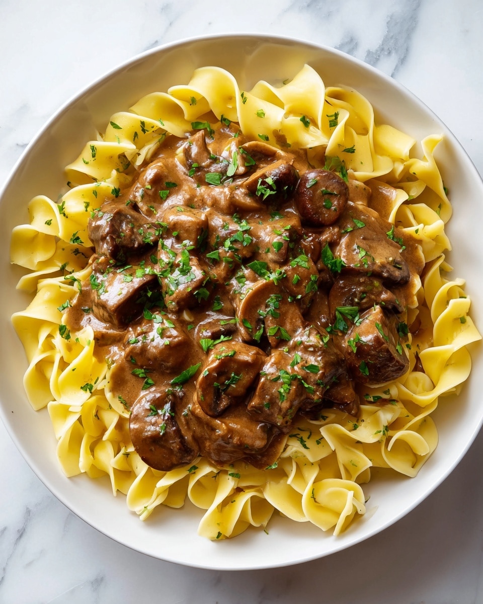 A white bowl filled with a base layer of curly, light yellow egg noodles. On top of the noodles, there is a thick layer of brown beef chunks and sliced mushrooms mixed in a creamy, light brown sauce with a smooth texture. Scattered over the dish are small pieces of fresh green parsley, adding a pop of color. The bowl is placed on a white marbled surface. photo taken with an iphone --ar 4:5 --v 7