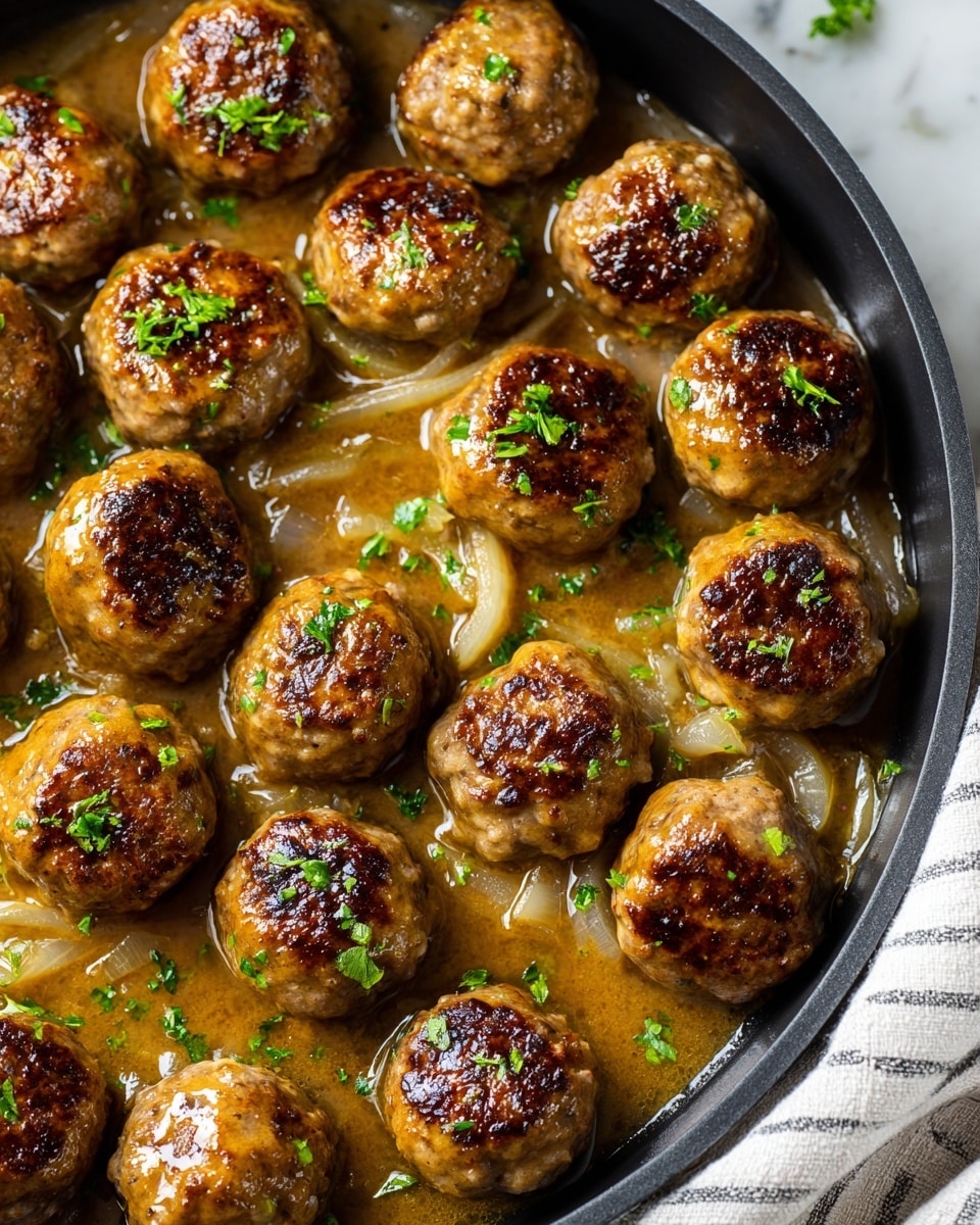 A round black skillet filled with about fifteen golden brown meatballs arranged closely together. Each meatball has a slightly shiny, glazed look with bits of cooked onions and browned spots on the surface. The meatballs sit in a glossy, light brown sauce with visible softened onion slices and small green herb pieces sprinkled on top. The skillet is placed on a white marbled surface with a white and gray striped cloth nearby, and a wooden dish with a yellow liquid and a wooden spoon is partly visible in the background. photo taken with an iphone --ar 4:5 --v 7