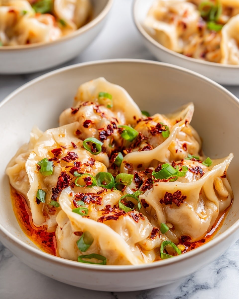 A white bowl filled with soft, pale dumplings covered in a shiny, spicy reddish-brown chili oil sauce that pools at the bottom. The dumplings have slightly wrinkled edges and a smooth texture, with chopped bright green scallions sprinkled evenly on top, adding a fresh contrast to the rich sauce. The bowl sits on a white marbled surface, with other similar bowls blurred in the background. Photo taken with an iphone --ar 4:5 --v 7