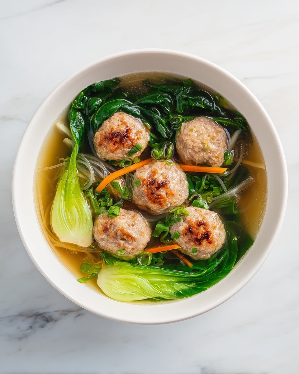 A white bowl filled with clear soup containing five light brown meatballs with a slightly browned top floating on the surface. Beneath the meatballs, there are several layers of bright green leafy vegetables and pieces of pale green bok choy, all submerged in the broth. Thin slices of orange carrot and white noodles are also visible beneath the meatballs, adding more color and texture to the dish. The bowl is placed on a white marbled surface. Photo taken with an iphone --ar 4:5 --v 7