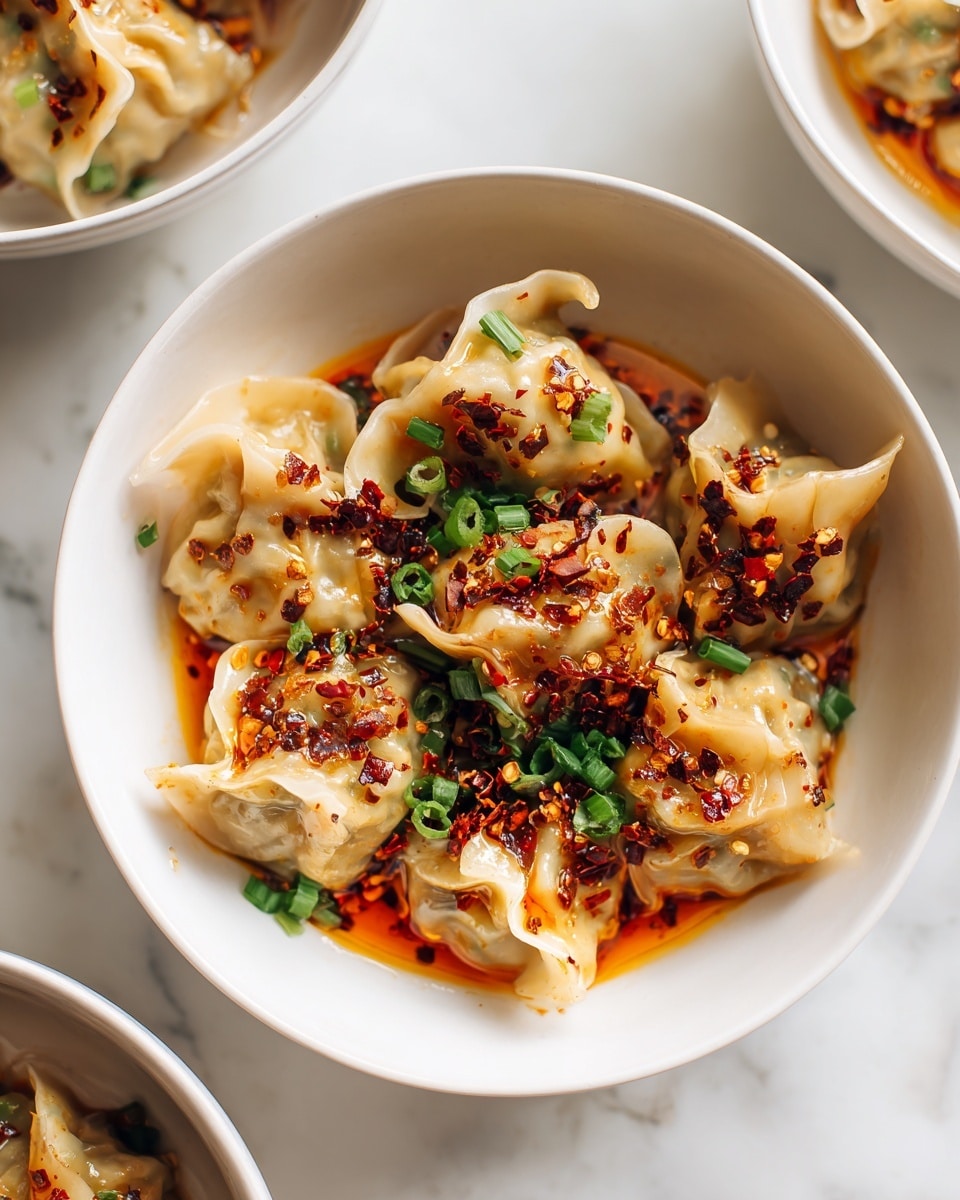 A white bowl filled with several light beige dumplings that have soft, slightly wrinkled textures. The dumplings are covered in a bright orange chili oil sauce that pools gently at the bottom of the bowl. On top, there are scattered pieces of deep red chili flakes and small bright green chopped spring onions, adding vibrant color contrast. The bowl sits on a white marbled surface with other similar bowls slightly out of focus in the background. Photo taken with an iphone --ar 4:5 --v 7