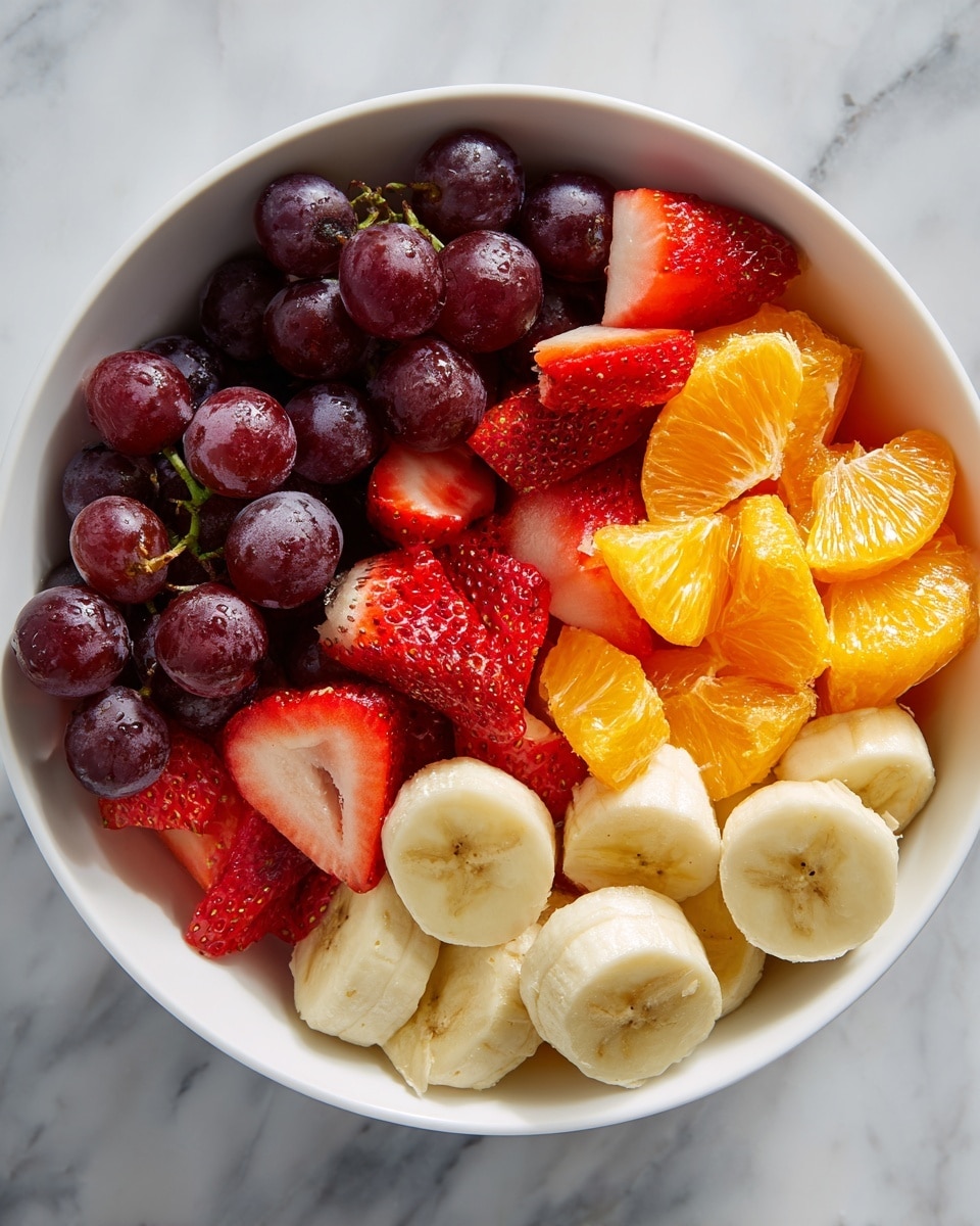 A white bowl filled with a colorful mix of fresh fruit pieces arranged in layers: at the bottom, thick slices of light yellow banana with visible seeds in the center; above them, bright orange chunks of juicy orange; scattered over the orange are halved strawberries showing their red color and small seeds; on one side, a cluster of shiny dark purple grapes still on small green stems; all fruit pieces have a fresh, slightly glossy texture suggesting juiciness. The bowl is placed on a white marbled surface. photo taken with an iphone --ar 4:5 --v 7